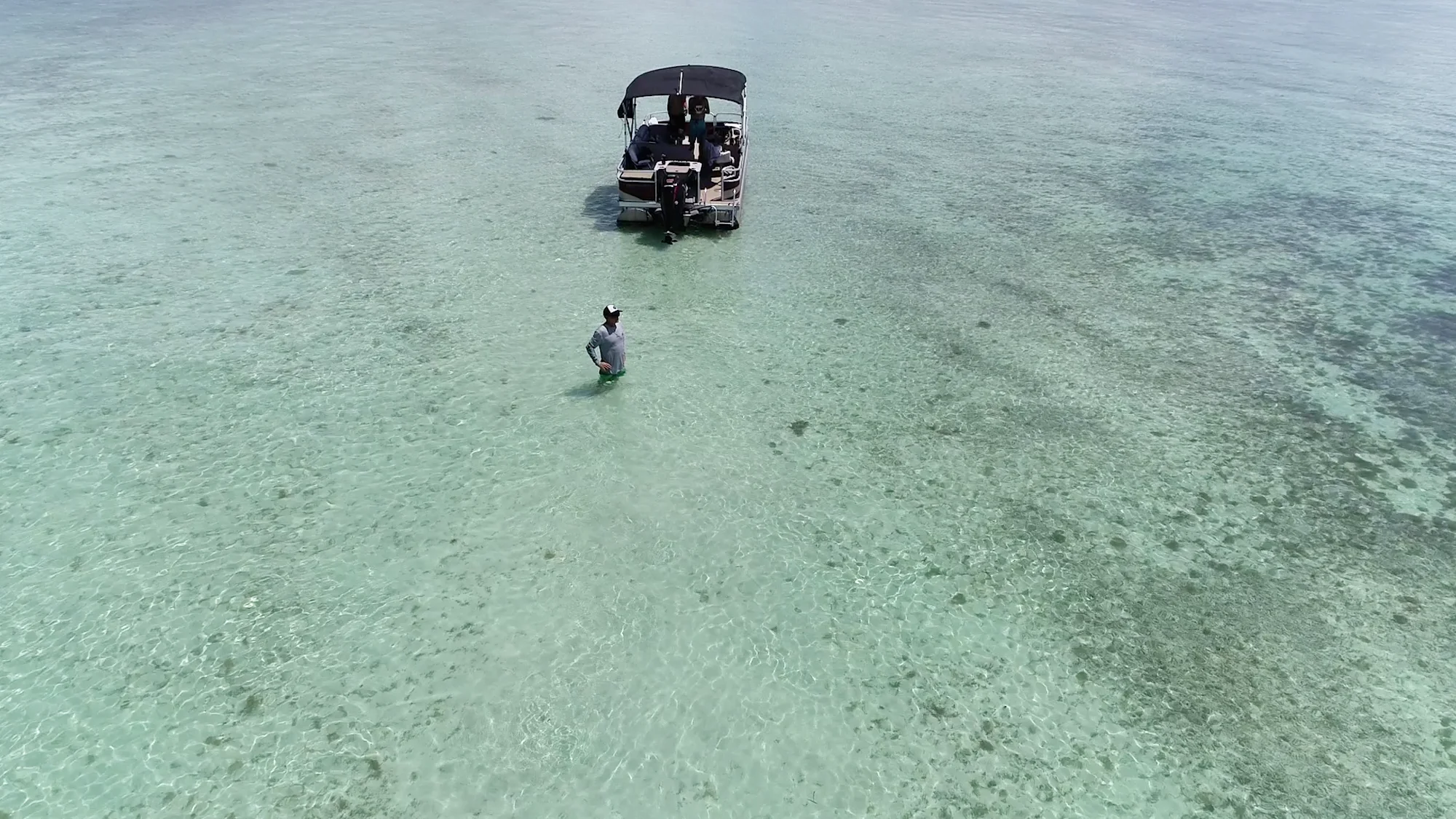 Aerial view of a pontoon boat anchored on a shallow turquoise sandbar, one person standing in the water