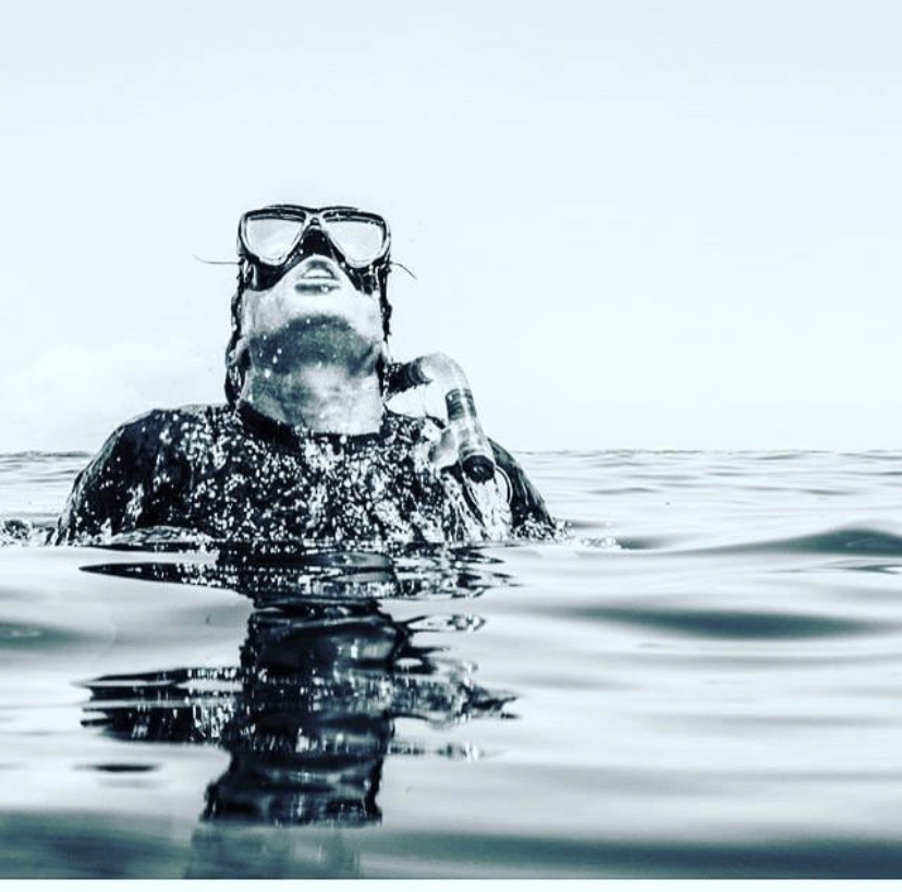 Black and white photo of a freediver surfacing with mask pushed up and a speargun in hand