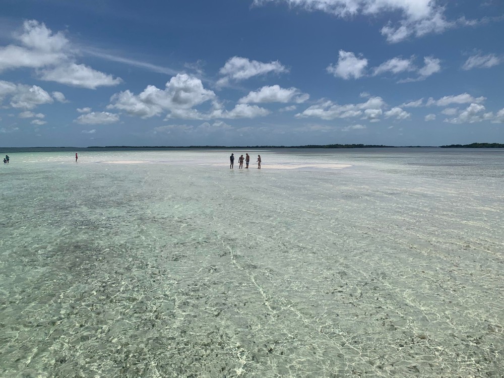 A group of people standing on an exposed sand spit surrounded by clear shallow water